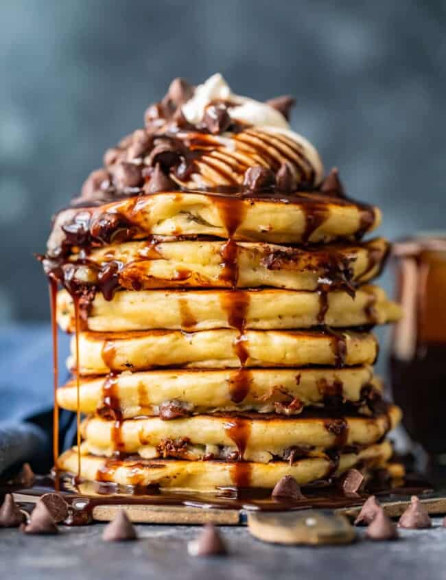 lots of chocolate chip pancakes stacked on each other drizzled with chocolate syrup and whipped cream