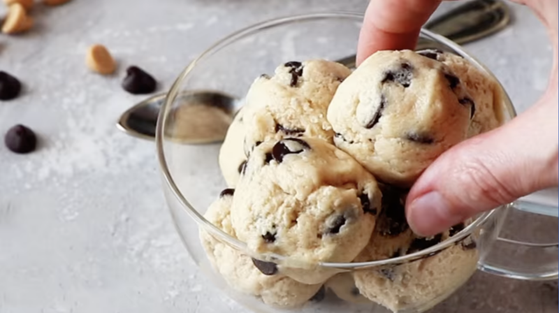 a hand grabbing an edible chocolate chip cookie dough ball from a glass bowl.