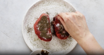 a hand seasoning filet mignon with salt and pepper on a white plate.