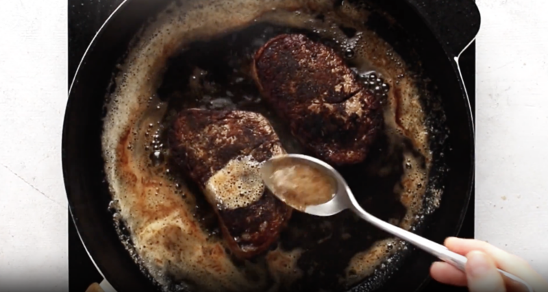 a spoon basting 2 filet mignon with butter in a cast iron skillet.