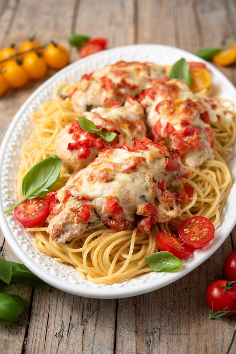 A close up of cheesy bruschetta chicken on a platter with spaghetti