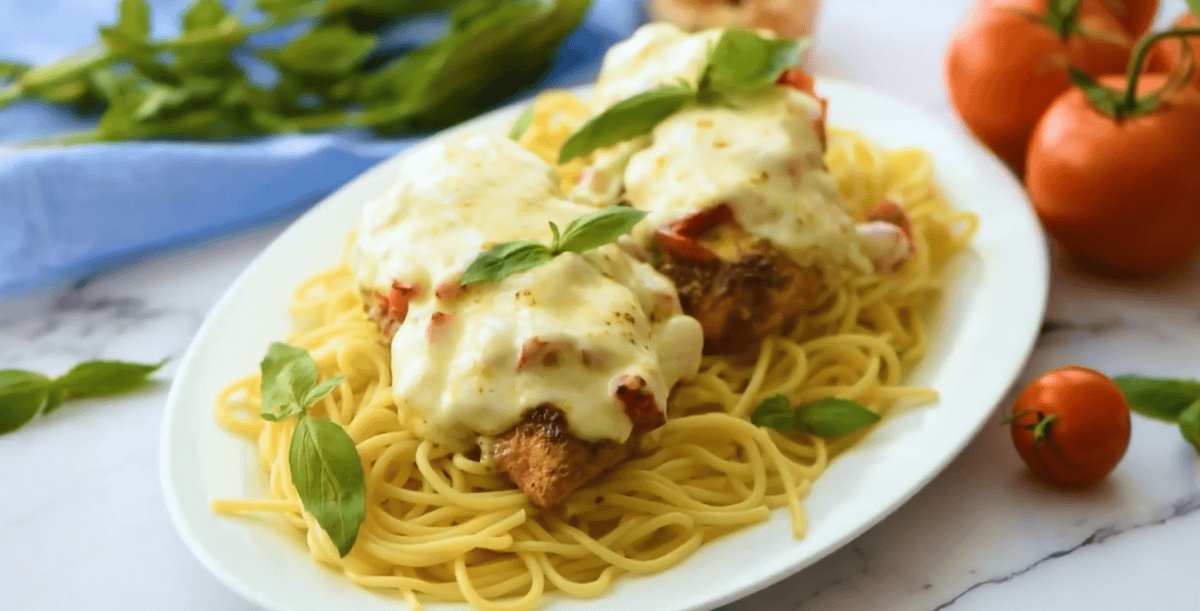 A serving of cheesy bruschetta chicken on top of spaghetti on a white plate. 