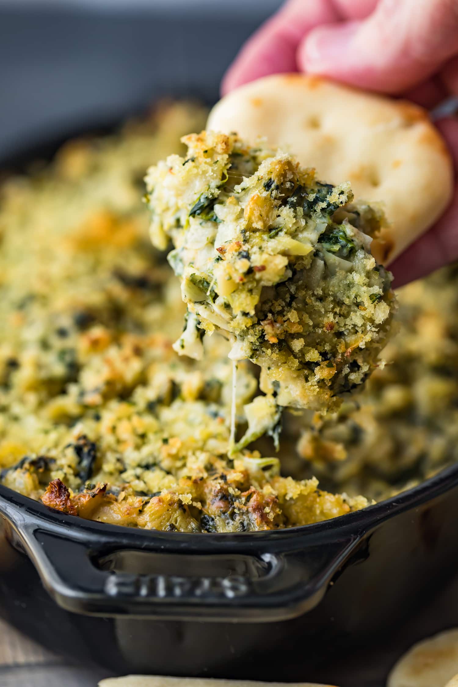 A person is dipping a cracker into a bowl of baked spinach and artichoke dip.