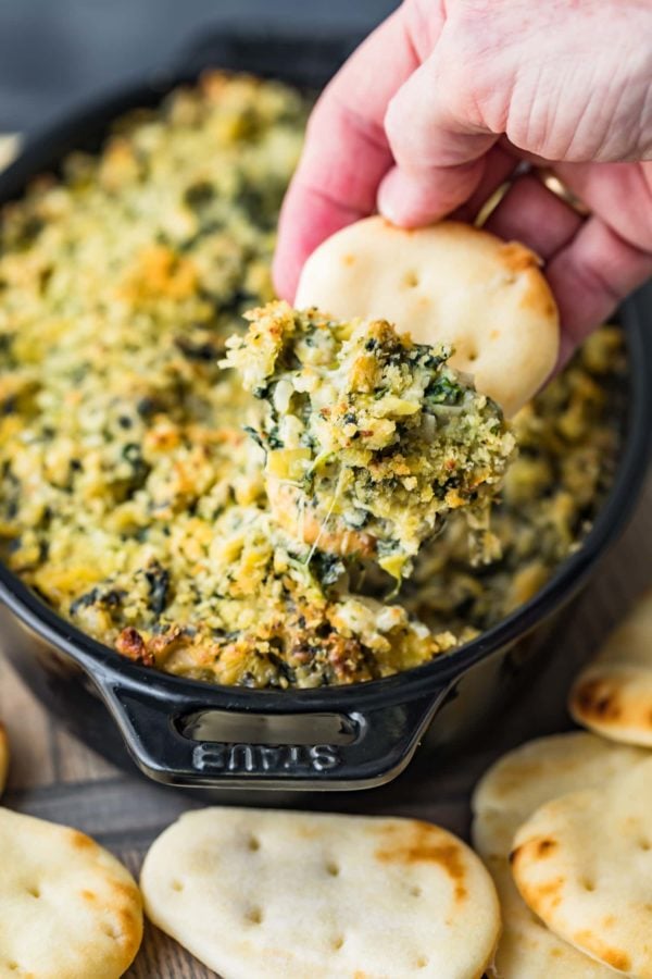 hand dipping a cracker into a dish of baked spinach dip.