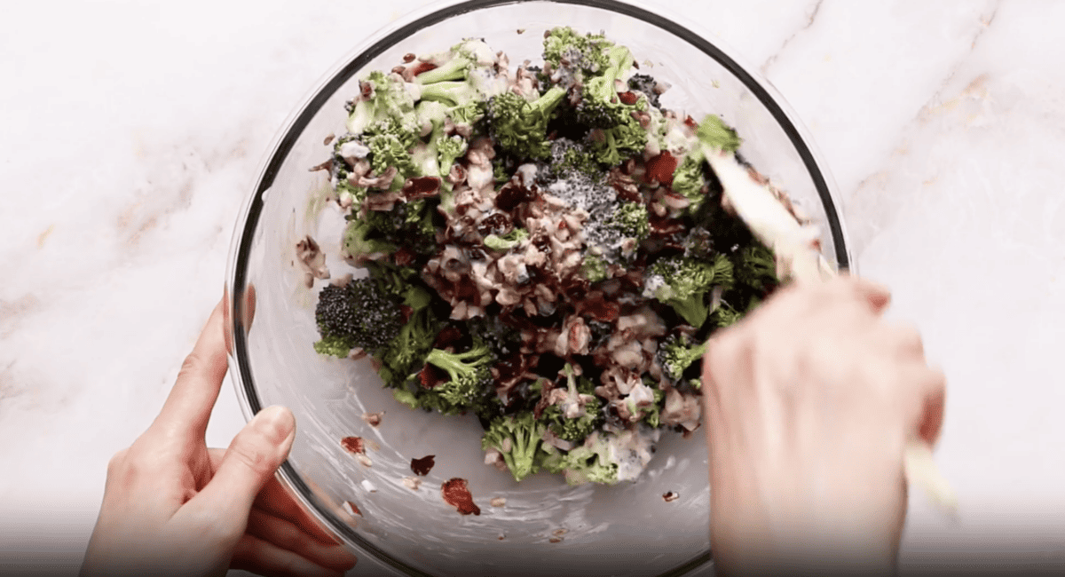 A person is mixing the broccoli salad in a bowl.