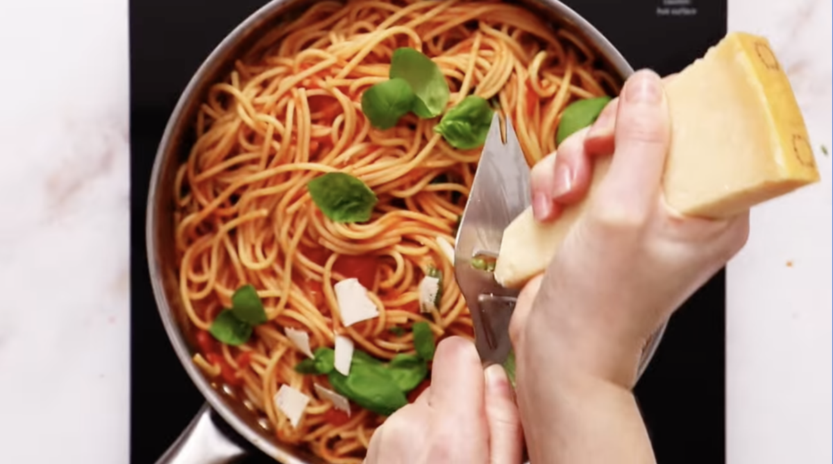 a person grating cheese over pasta pomodoro in a pan.