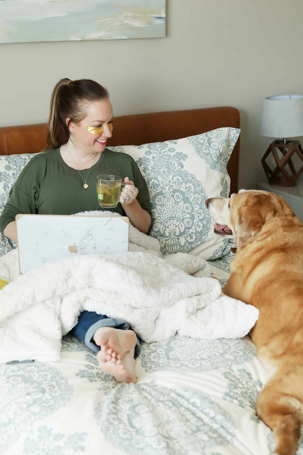woman in bed drinking tea smiling at dog