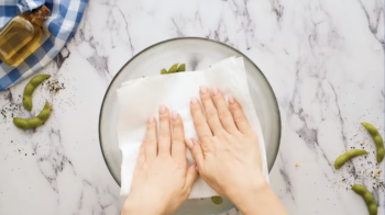 hands pressing a paper towel into cooked edamame in a glass bowl.