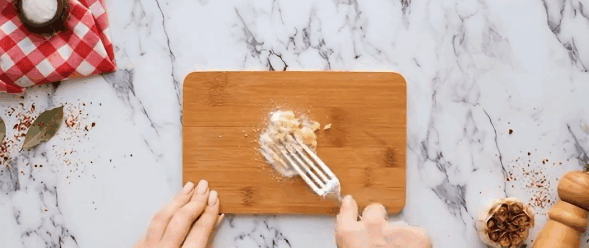 Preparing garlic paste on a wooden cutting board.