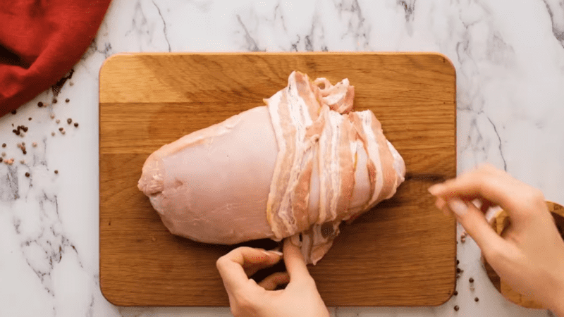 turkey breast sitting on a cutting board, hands wrapping turkey with slices of bacon