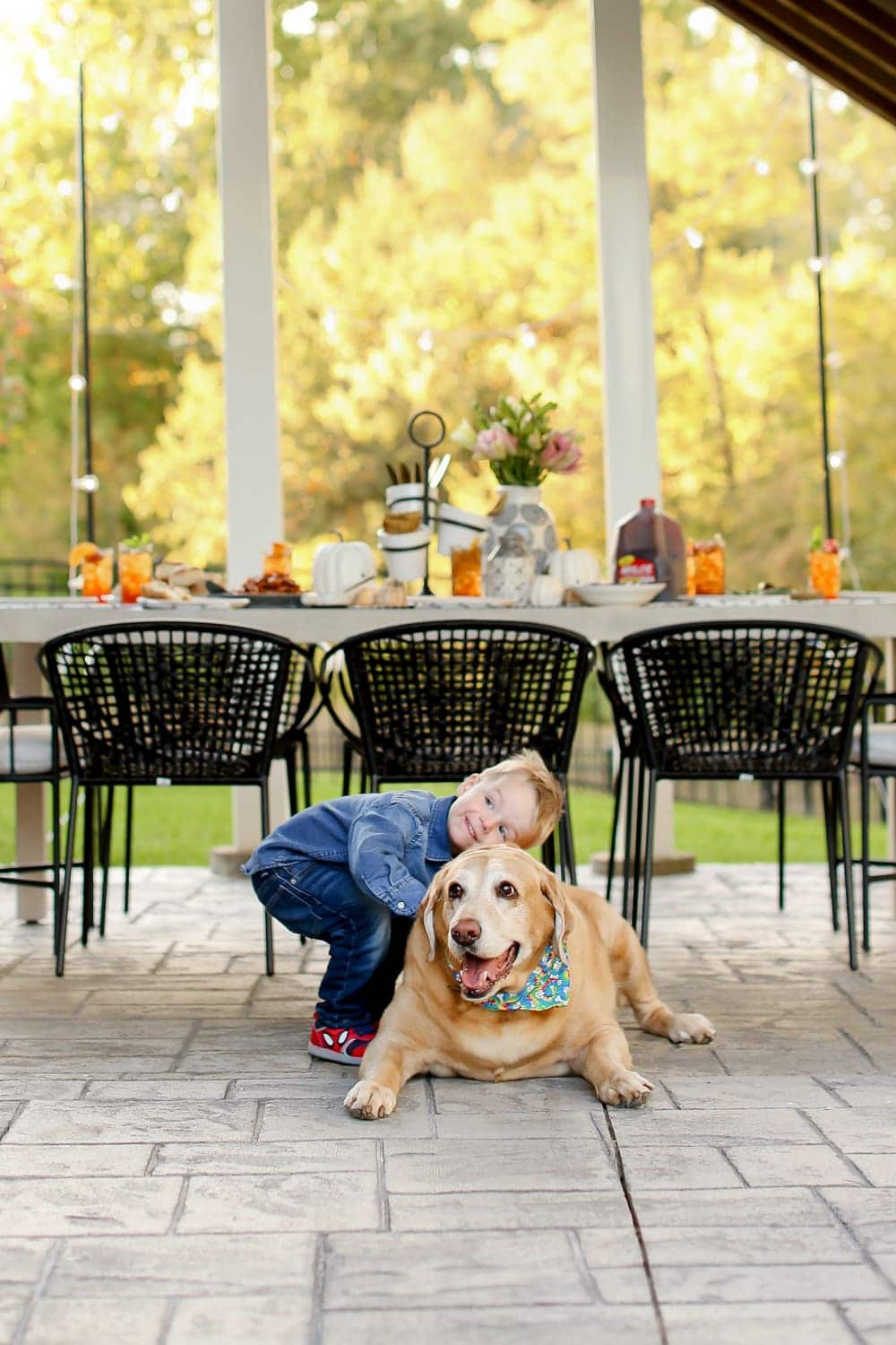 A toddler boy hugging a yellow labrador retriever with a table behind them.