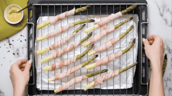 uncooked bacon wrapped asparagus lined up on a wire rack set in a baking sheet.