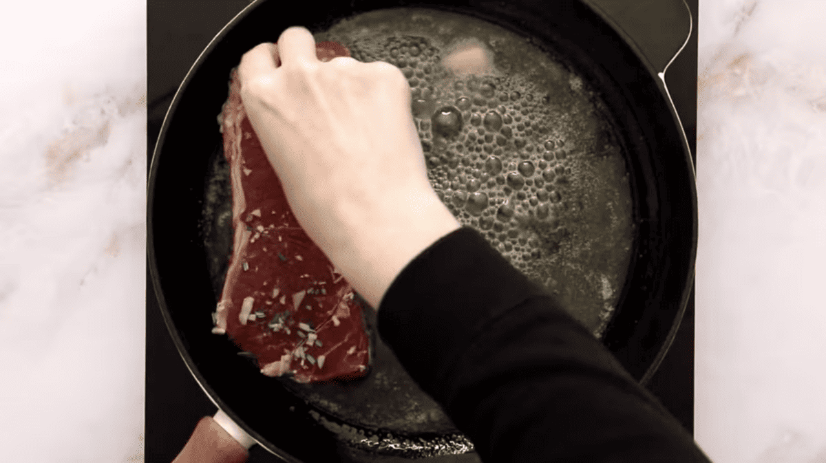 A person is frying a rosemary steak in a pan.