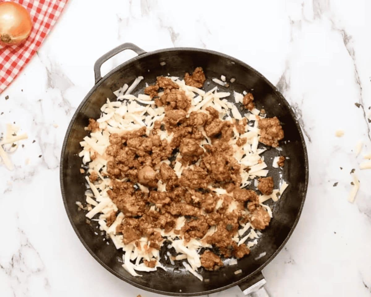 First layer of shredded cheese topped with browned chorizo in a cast iron skillet.