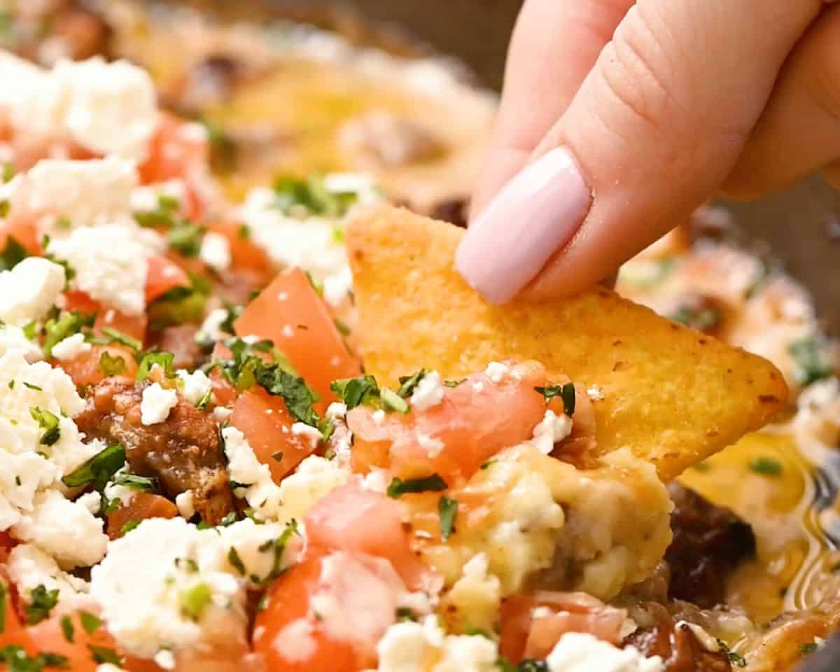 Tortilla chip scooping cheesy queso fundido topped with chorizo, tomatoes, cilantro, and queso fresco.