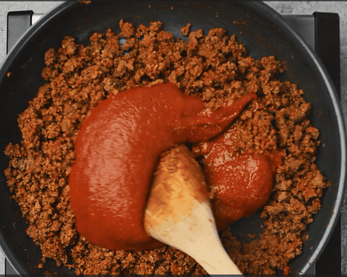 Cooked ground beef with tomato sauce being stirred in.