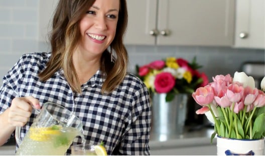 a woman holding a pitcher of lemonade in front of flowers.