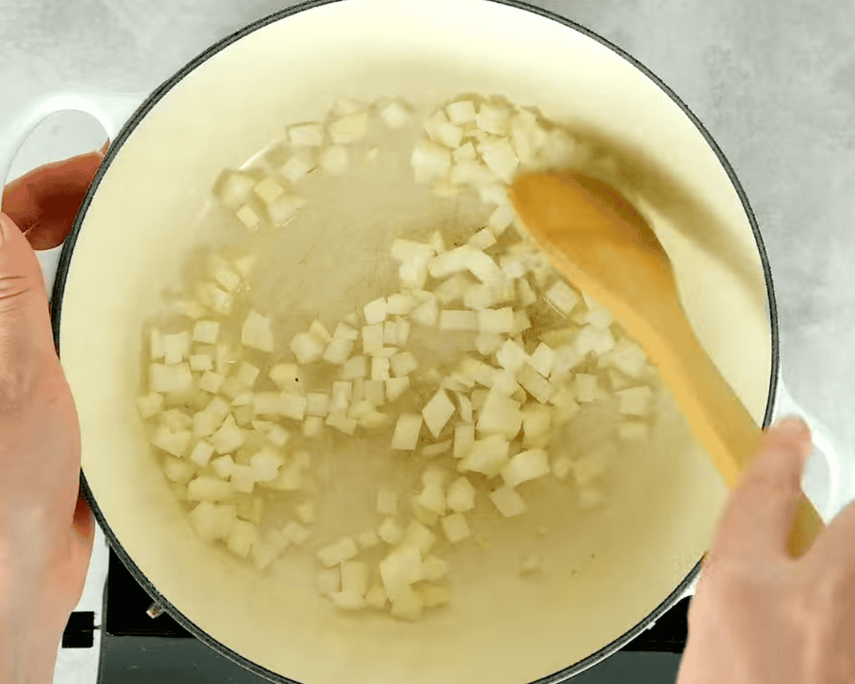 Chopped onions sautéing in a white Dutch oven with oil, being stirred with a wooden spoon.