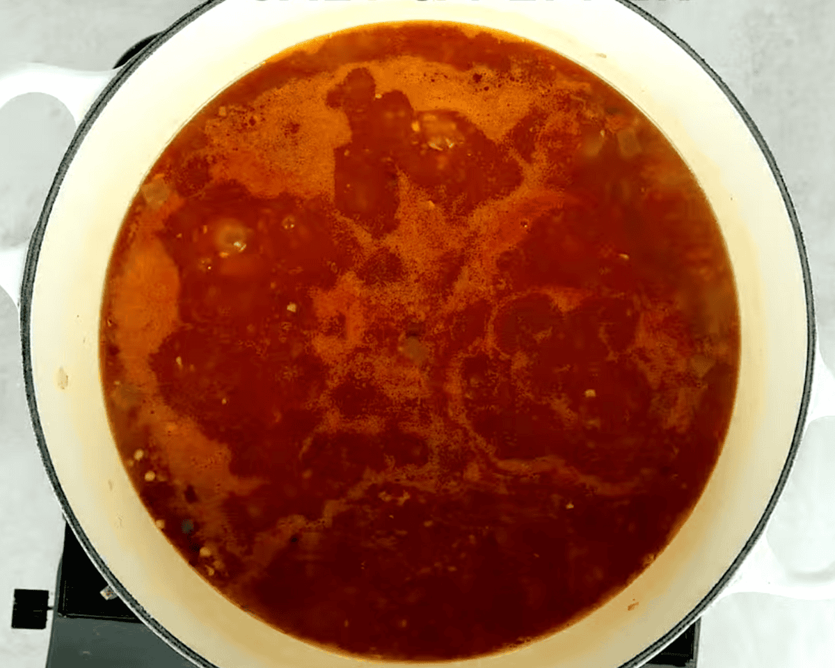 A simmering tomato-based broth in a white Dutch oven, rich in color with small bubbles forming on the surface.