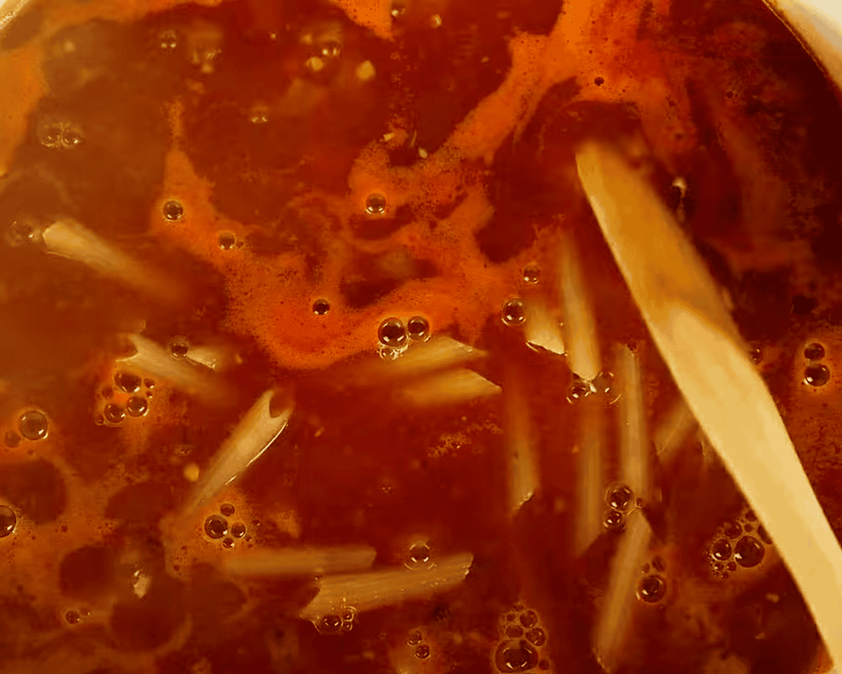 Uncooked penne pasta being stirred into the simmering red broth with a wooden spoon.