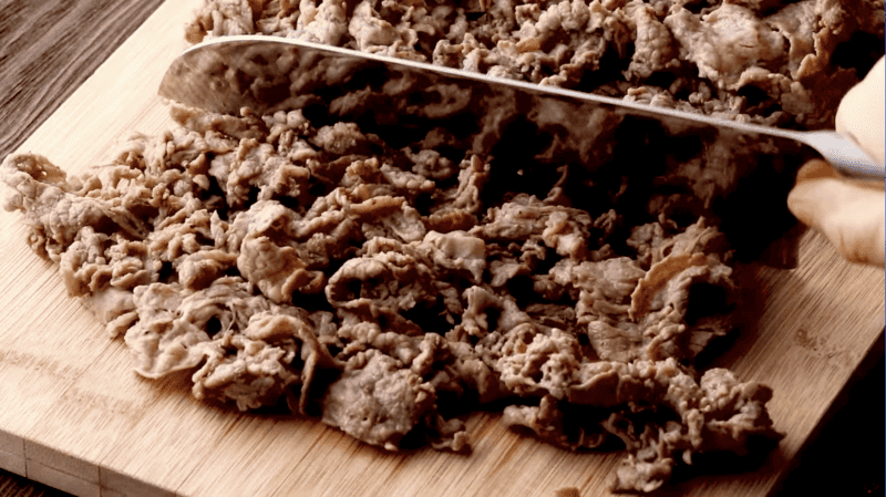 A person preparing a philly cheesesteak dip by cutting meat on a cutting board.