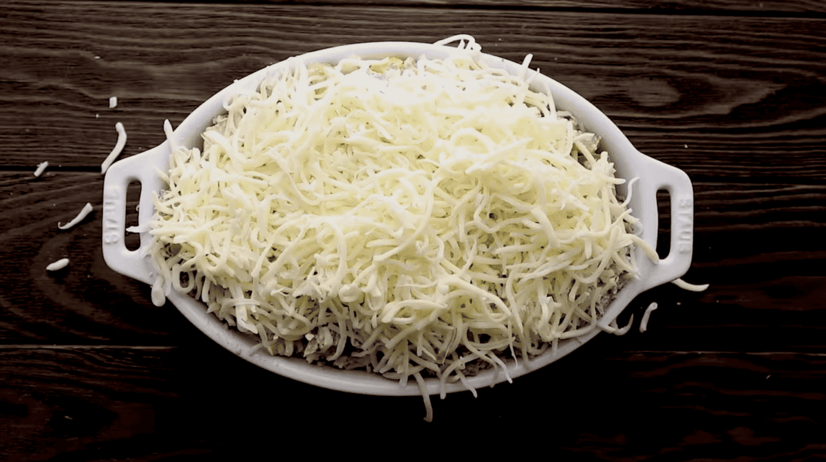 Philly cheesesteak dip with shredded cheese in a white dish on top of a wooden table.