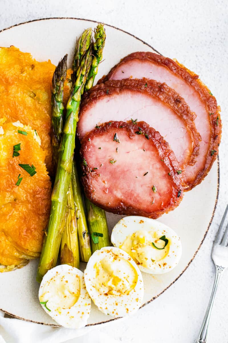 A dinner plate filled with slices of ham, stalks of asparagus, scoops of potatoes, and hard-boiled eggs.