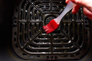 a red silicone pastry brush brushing oil on the basket of an air fryer.