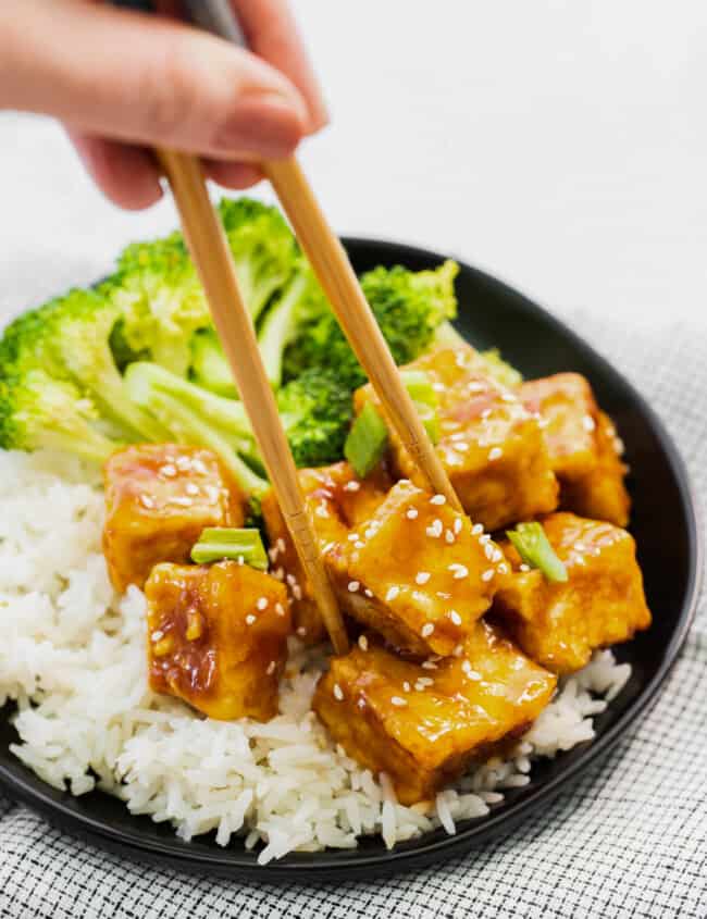 chopsticks grabbing a cube of air fryer teriyaki tofu on a black plate.