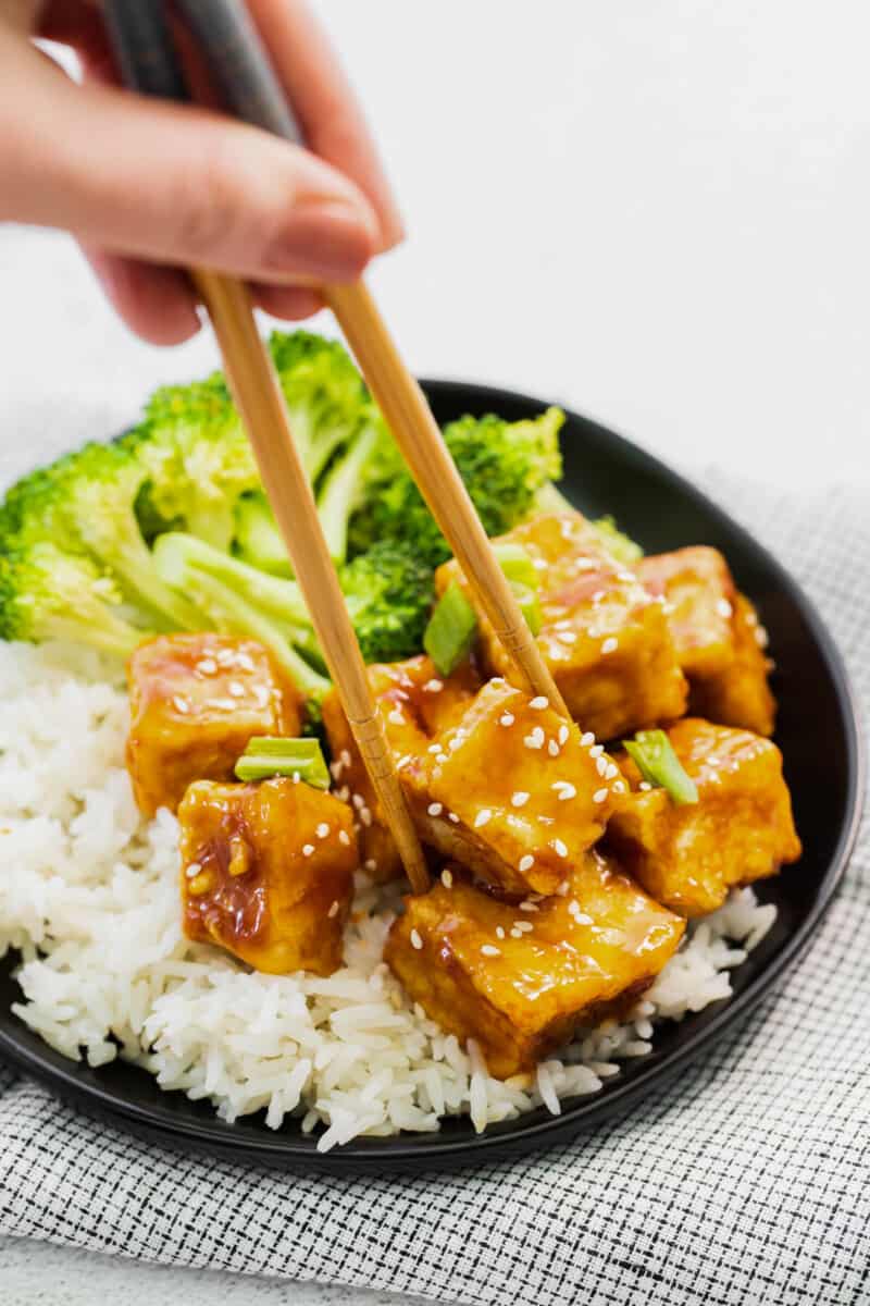 chopsticks grabbing a cube of air fryer teriyaki tofu on a black plate.