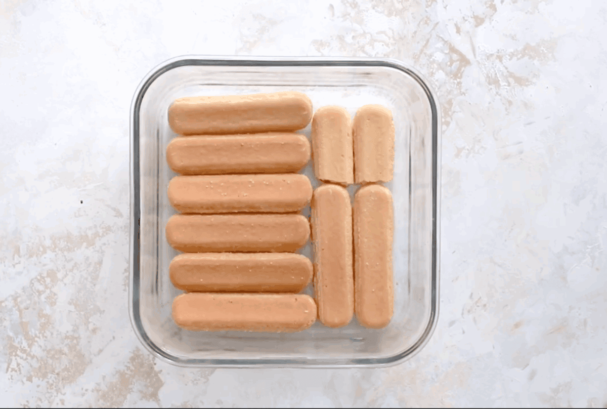 A 9x9-inch glass baking dish with a bottom layer of 12 soft ladyfingers arranged in rows to form the base layer of the tiramisu.