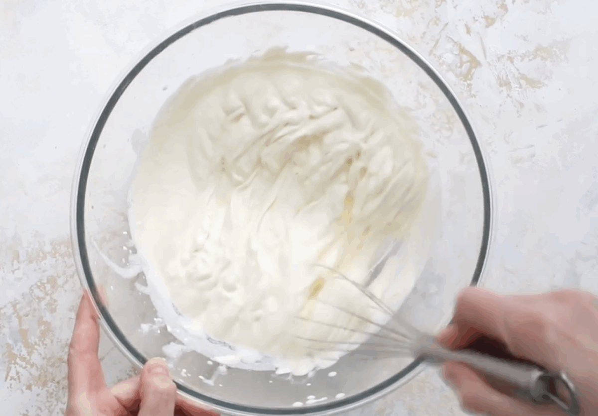A large glass bowl filled with soft peaks of whipped cream, being whisked by hand to incorporate air and build volume for the tiramisu filling.