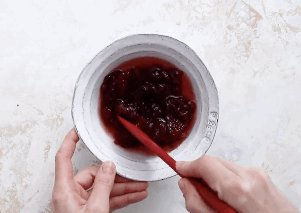 A small white bowl filled with strawberry jam mixed with raspberry liqueur, being stirred with a red spatula.