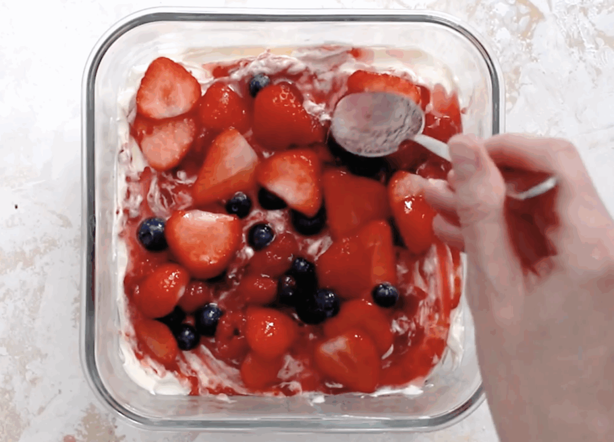 A hand spooning the berry mixture over the cream layer, with fresh strawberries, raspberries, and blueberries visibly spread across the surface.