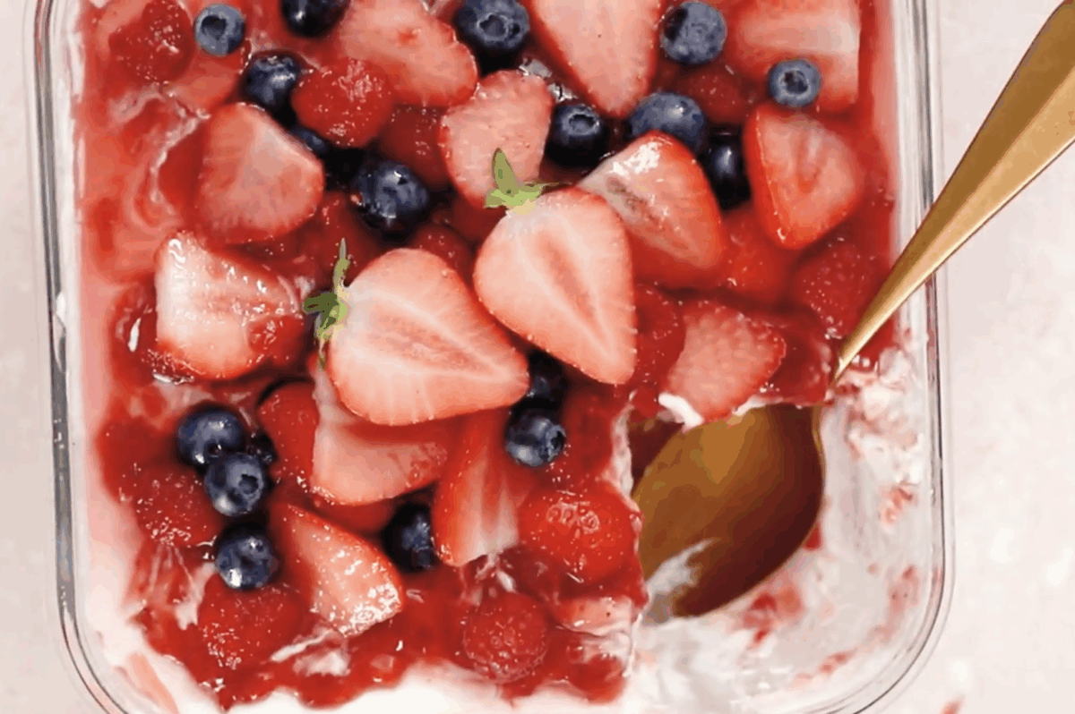 A close-up of a glass dish of strawberry tiramisu with a golden spoon scooping out a portion, revealing layers of mascarpone cream, ladyfingers, and a vibrant topping of fresh strawberries, raspberries, and blueberries.