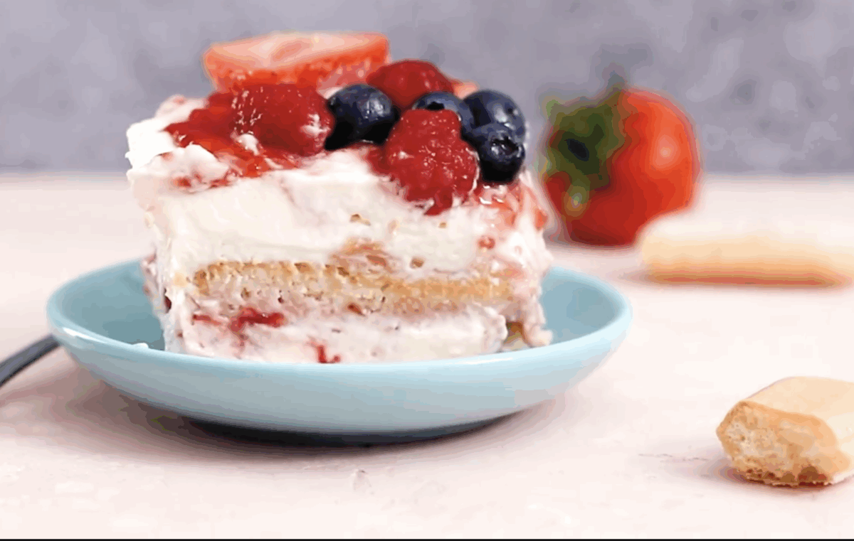 A single slice of strawberry tiramisu served on a light blue plate, showing distinct layers of creamy filling, soaked ladyfingers, and a colorful berry topping with whole raspberries and blueberries.

