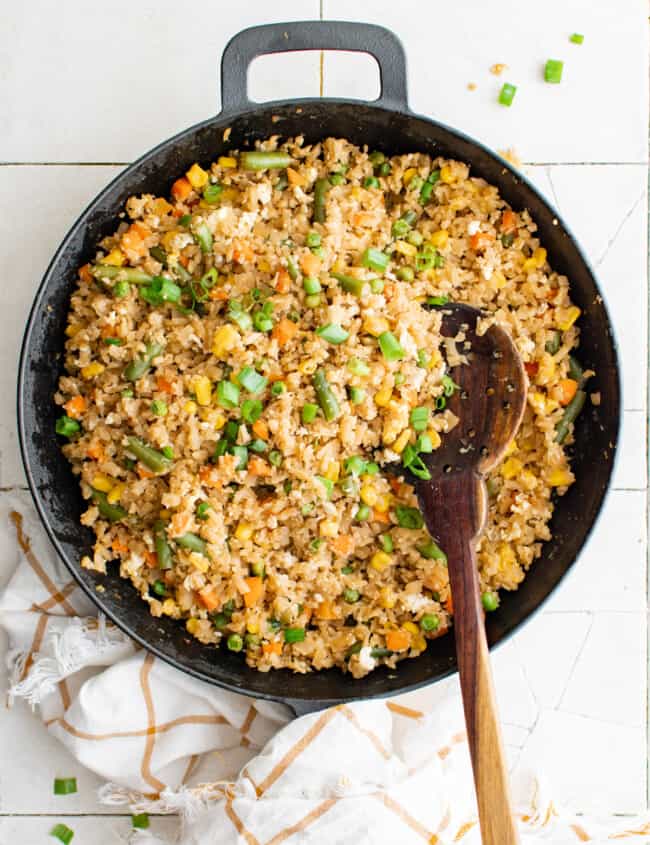 overhead view of cauliflower fried rice in a cast iron pan with a wooden spoon.