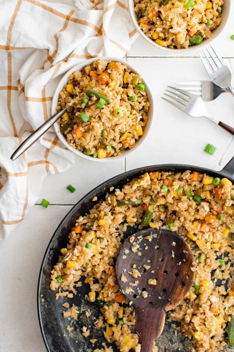 cauliflower fried rice in a white bowl with a fork next to a pan of rice.