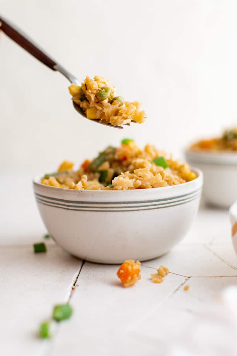 a forkful of cauliflower fried rice suspended over a white bowl filled with cauliflower fried rice.