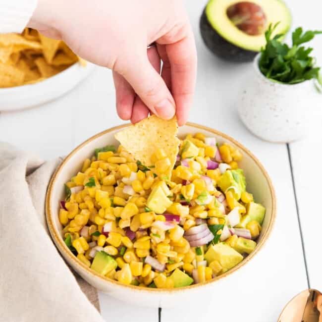 a hand dipping a chip into corn and avocado salsa in a white bowl.