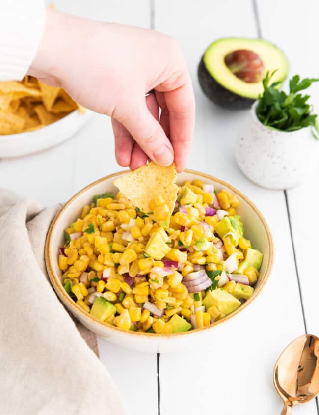 a hand dipping a chip into corn and avocado salsa in a white bowl.