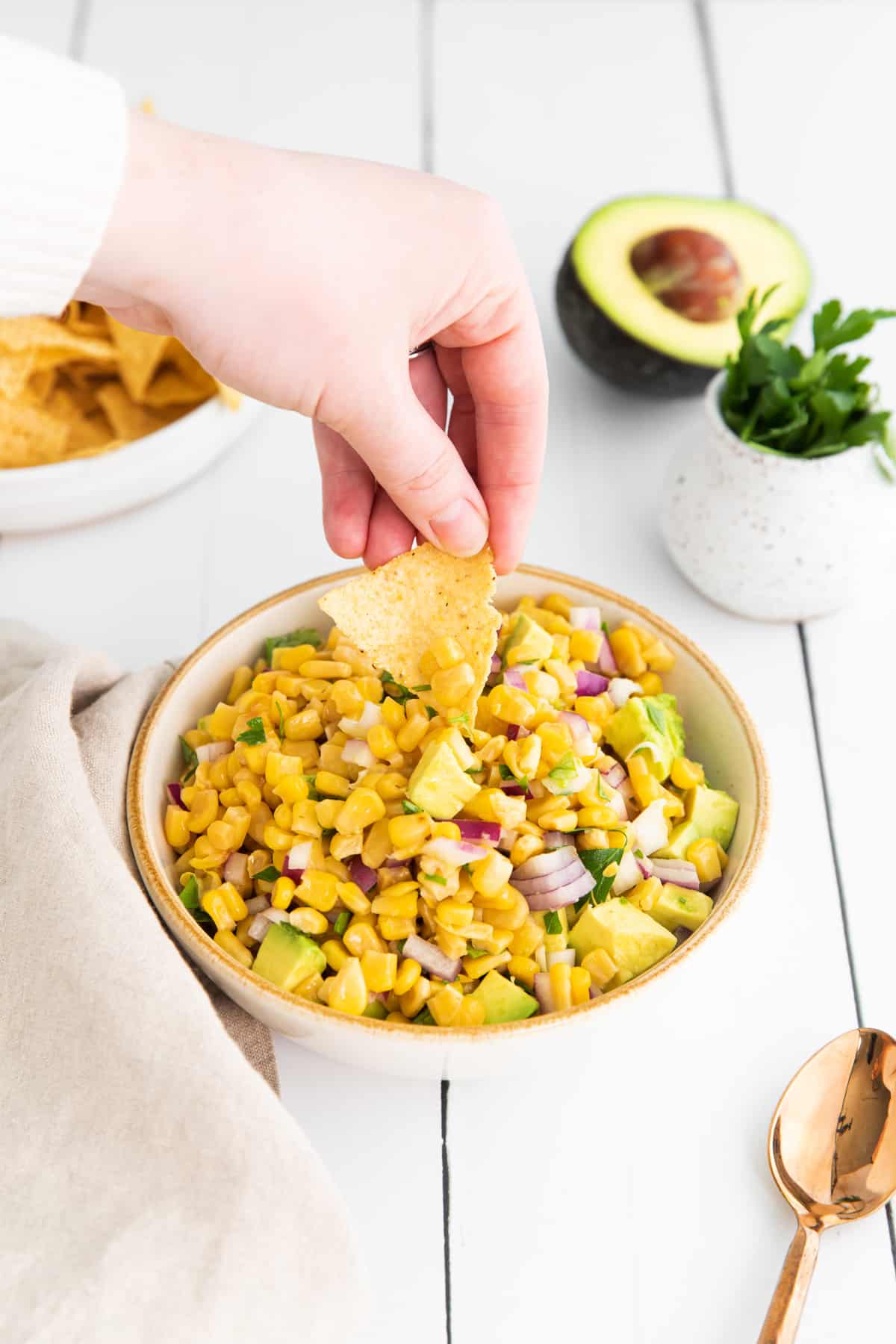 a hand dipping a chip into corn and avocado salsa in a white bowl.