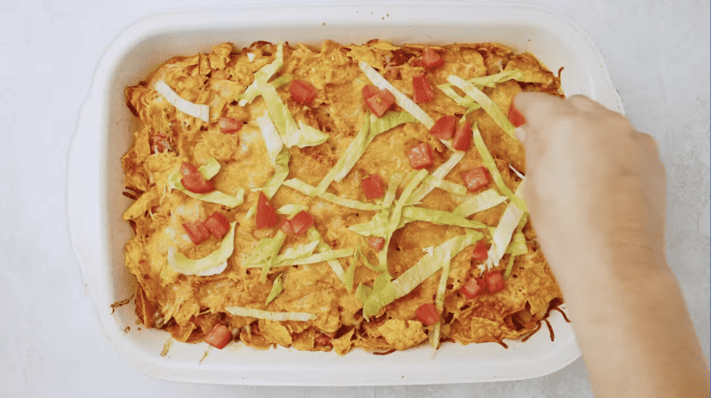 A person reaches into a Doritos casserole dish to add toppings.