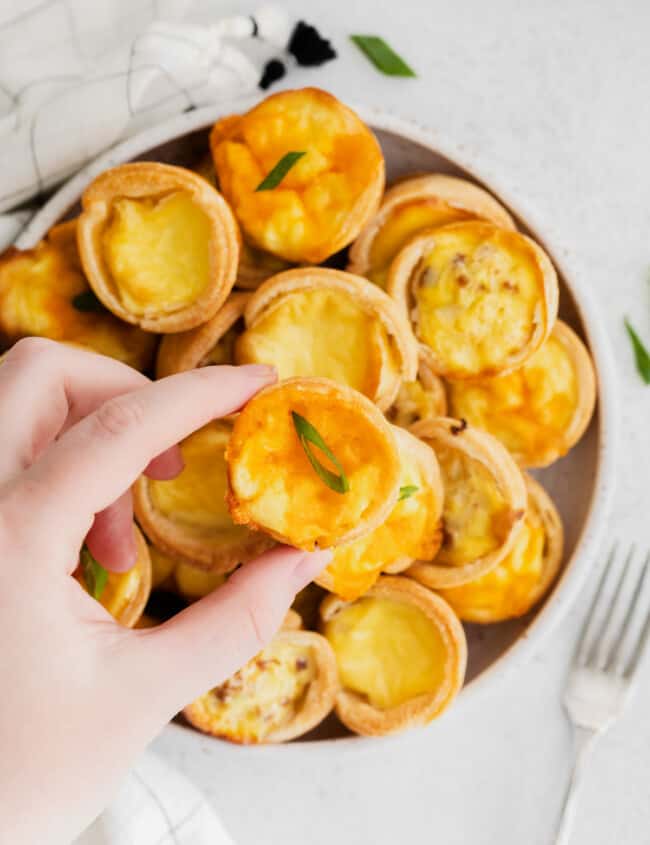 hand grabbing a mini quiche from a white plate of mini quiches.