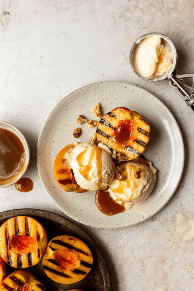 grilled peaches with ice cream and caramel sauce on a plate with a side of caramel sauce and more grilled peaches.