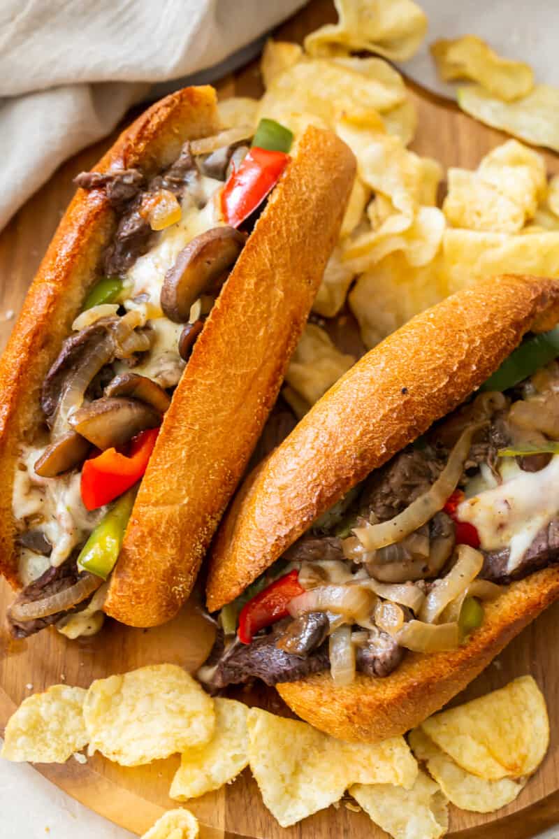 two philly cheesesteaks on cutting board with chips