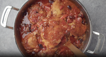 overhead view of a chicken thigh on a wooden spoon above chicken cacciatore in a pan.