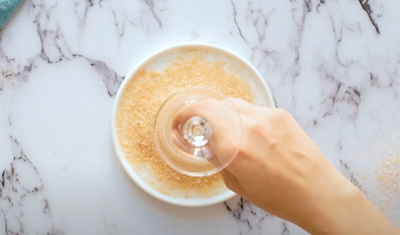 hand holding a champagne flute upside down and dipping the rim into gold sprinkles on a white plate