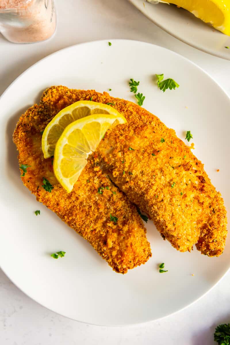 breaded tilapia filet topped with lemon slices on a white plate