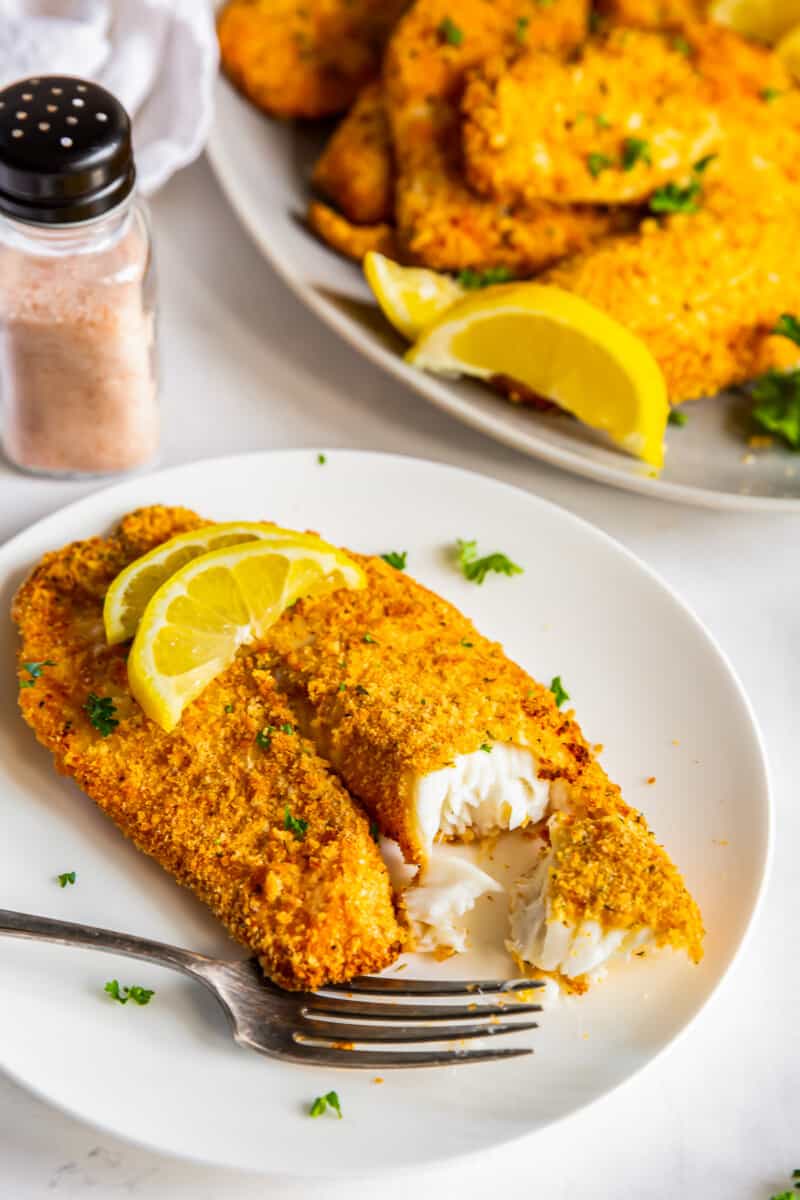 bite out of breaded tilapia filet topped with lemon slices on a white plate with a fork
